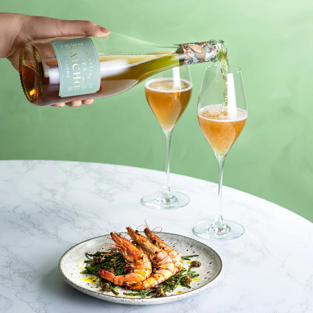 Person pouring Saicho Hojicha Sparkling Tea into two glasses with a plate of shrimp on a marble surface.