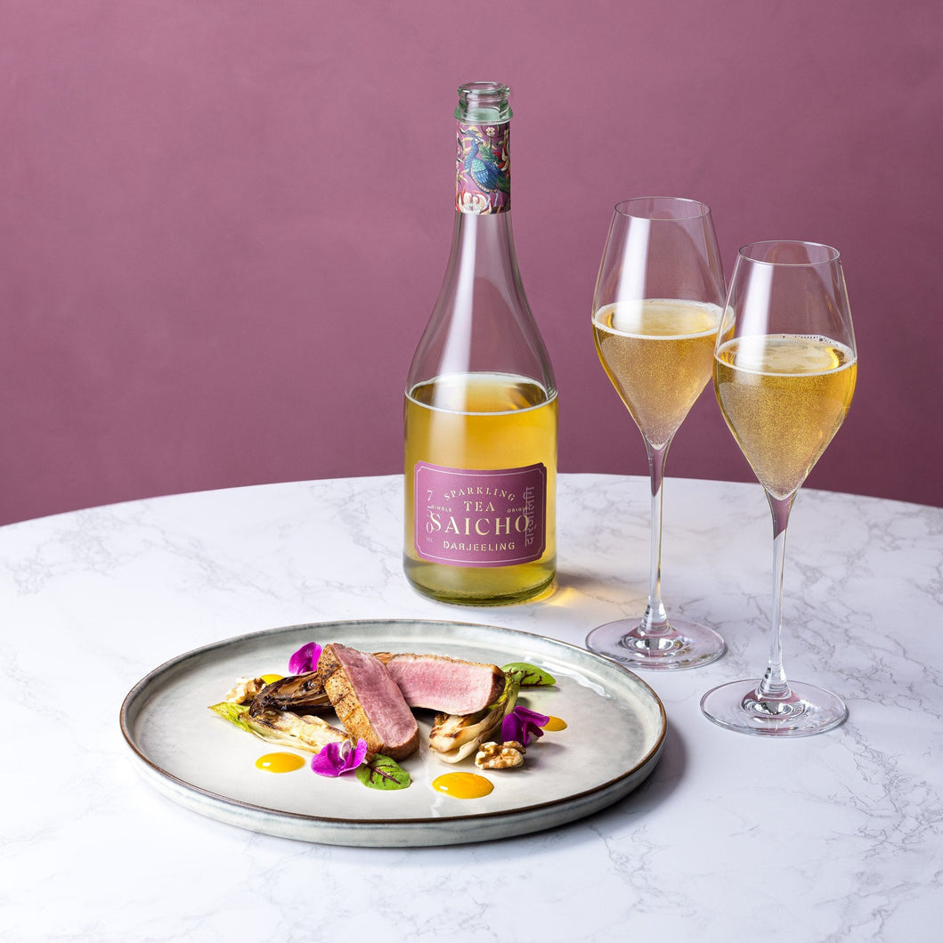 Bottle of Saicho Darjeeling Sparking Tea, two glasses with wine, and a dish of food on a marble surface with a purple background